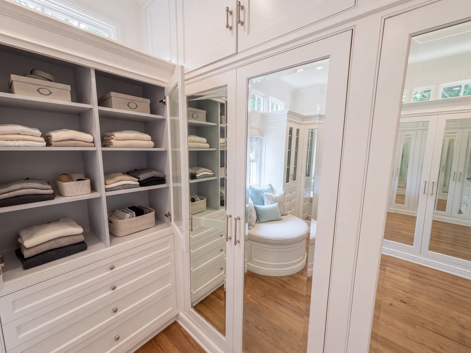 Primary closet with white custom millwork, glass-front uppers, mirrored doors and a built-in window seat
