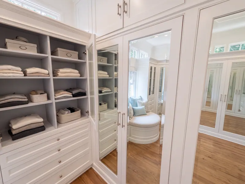 Primary closet with white custom millwork, glass-front uppers, mirrored doors and a built-in window seat