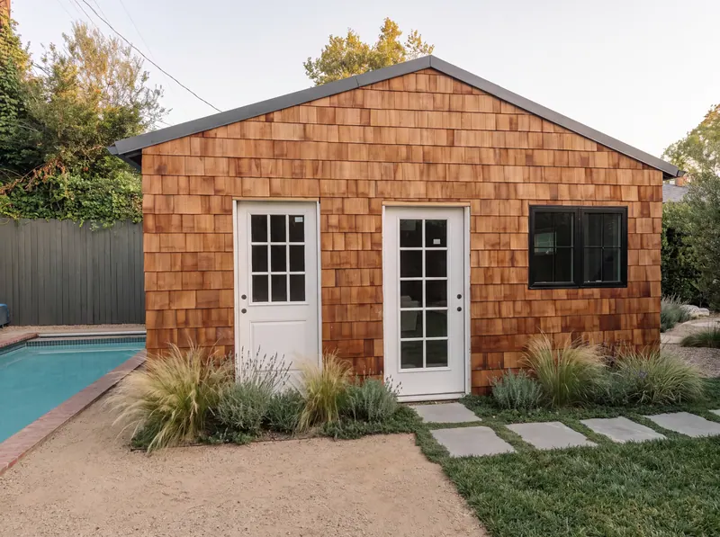 Detached backyard studio clad in cedar shingles, with glass-paned doors, a black-framed window and drought-tolerant planting by the pool