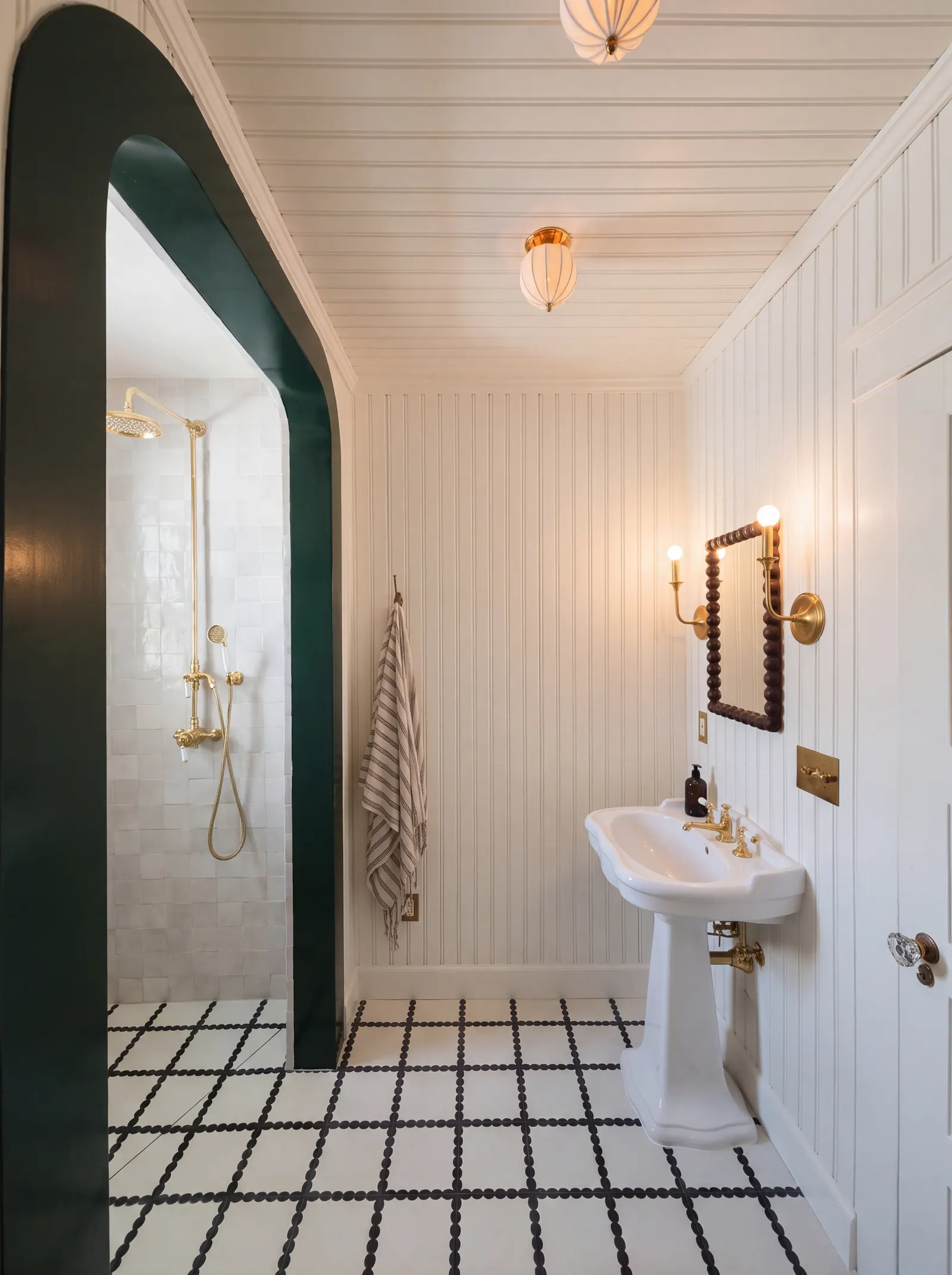 Craftsman bath with beadboard walls, a green lacquered arch to a white zellige shower, brass shower column, pedestal sink and bobbin mirror