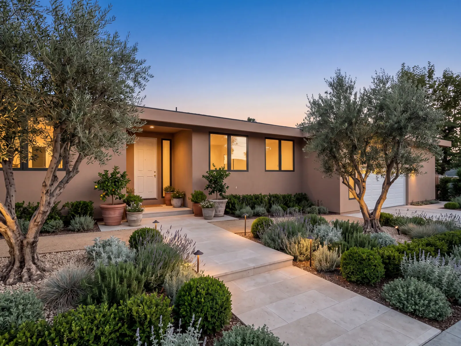 Single-story home at dusk, with sand-toned stucco, dark window frames, olive trees and a stone walk