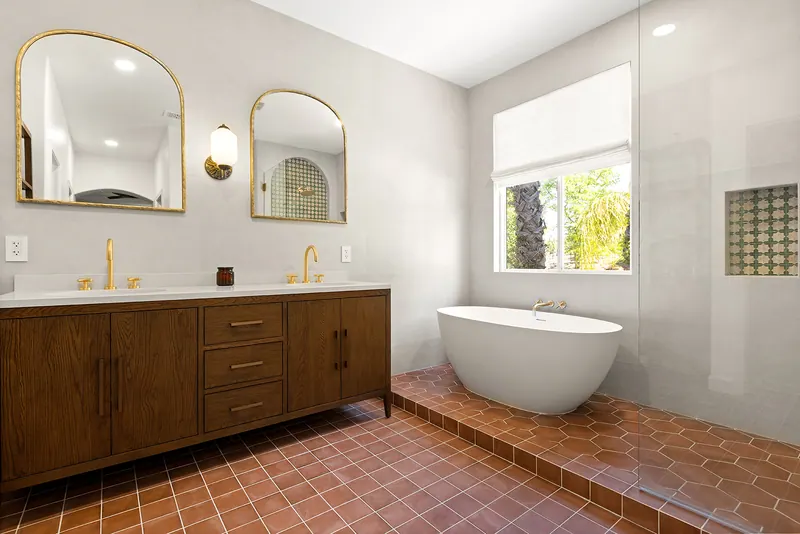 Walnut-tone double vanity with arched brass mirrors and a freestanding tub