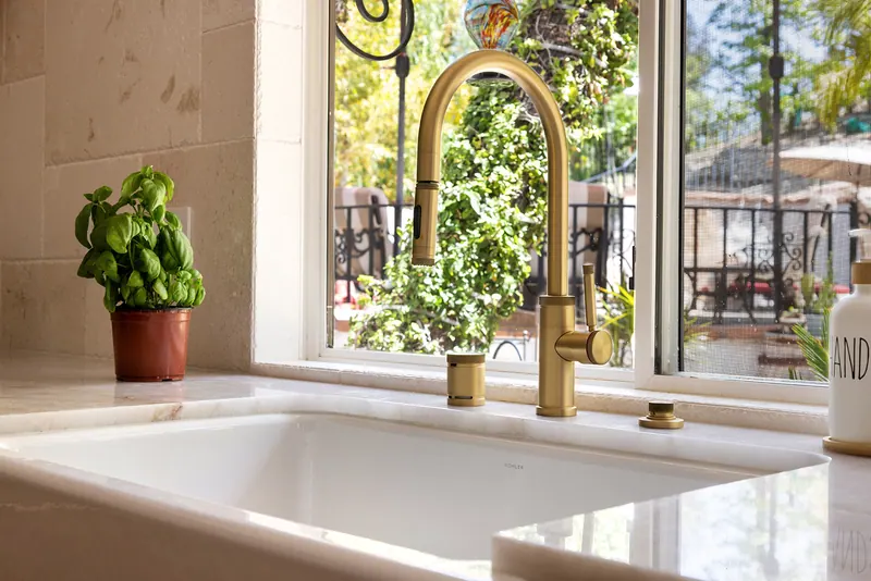 Brass faucet over a white farmhouse sink below the window