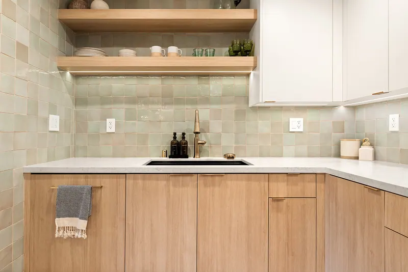 Sink wall with glazed sage tile, floating oak shelves and a brass faucet